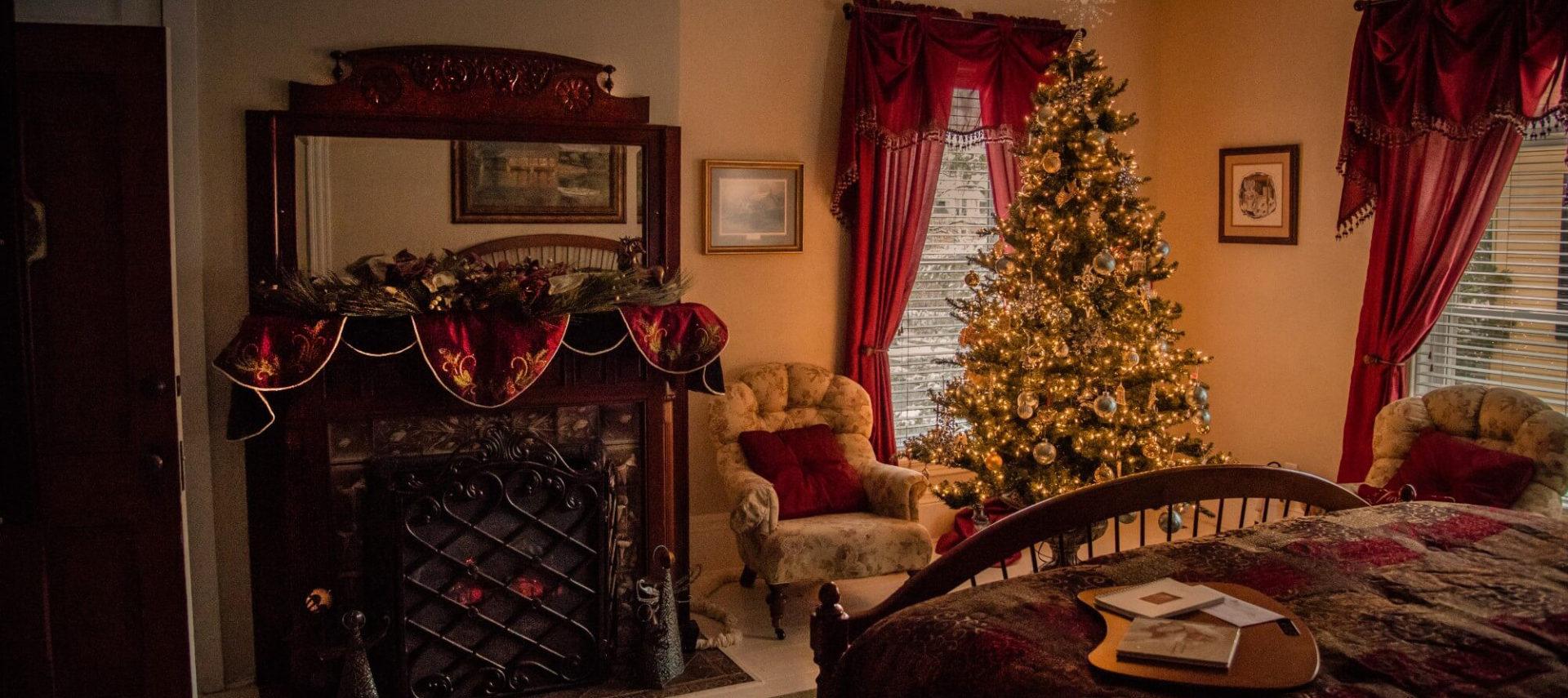 King bed with wheat footboard, burgundy bedding and view of vintage tiled fireplace with beige print upholstered chairs and Christmas tree