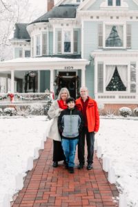 Innkeeper mom dad and son pose in front of snowy blue Victorian home