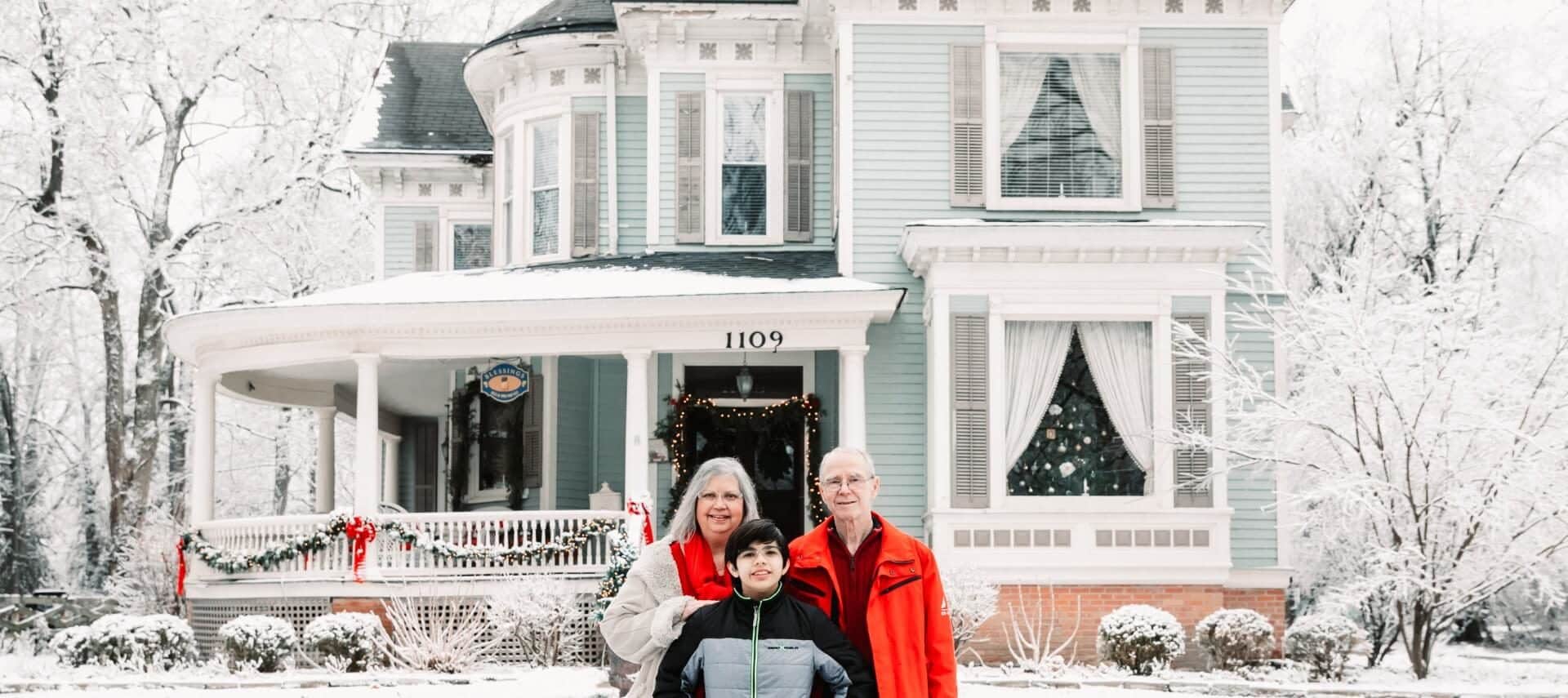 Innkeeper mom dad and son pose in front of snowy blue Victorian home with white columned porch