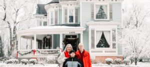Innkeeper mom dad and son pose in front of snowy blue Victorian home with white columned porch
