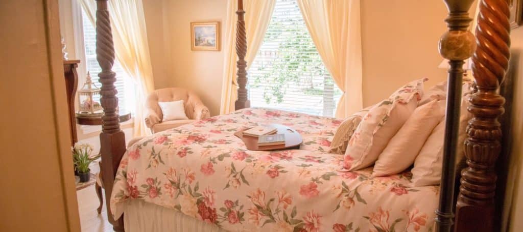 Queen size four poaster bed with colorful bedding with windows and a chair in the background