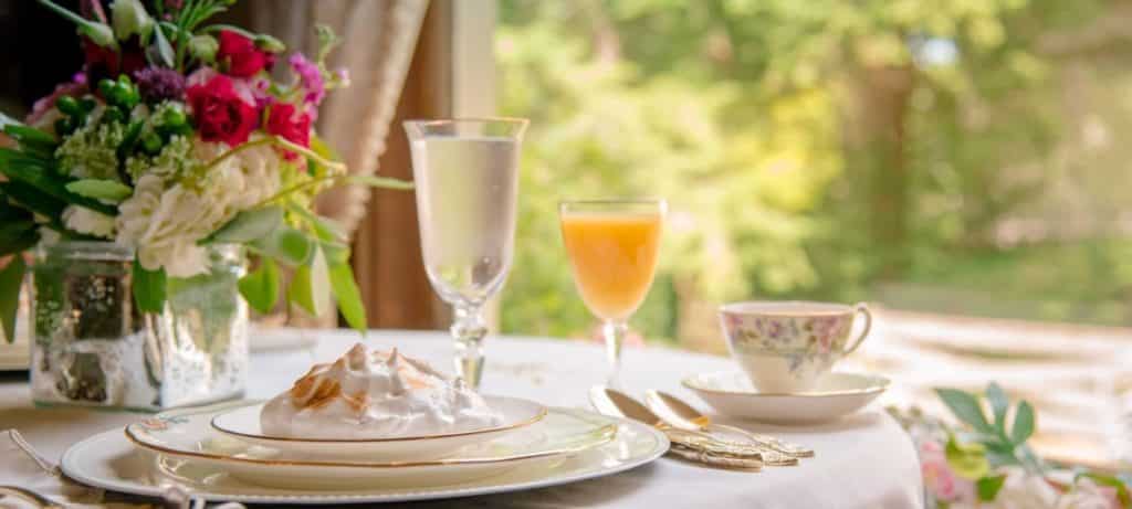 Tabletop with toasted meringue dessert on fine china plates crystal glasses of water and juice and colorful floral arrangement