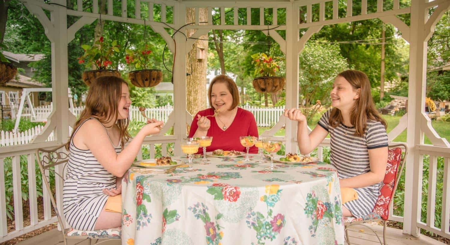 Lady and two girls sitting at a table under a gazeebo eating, smiling and laughing