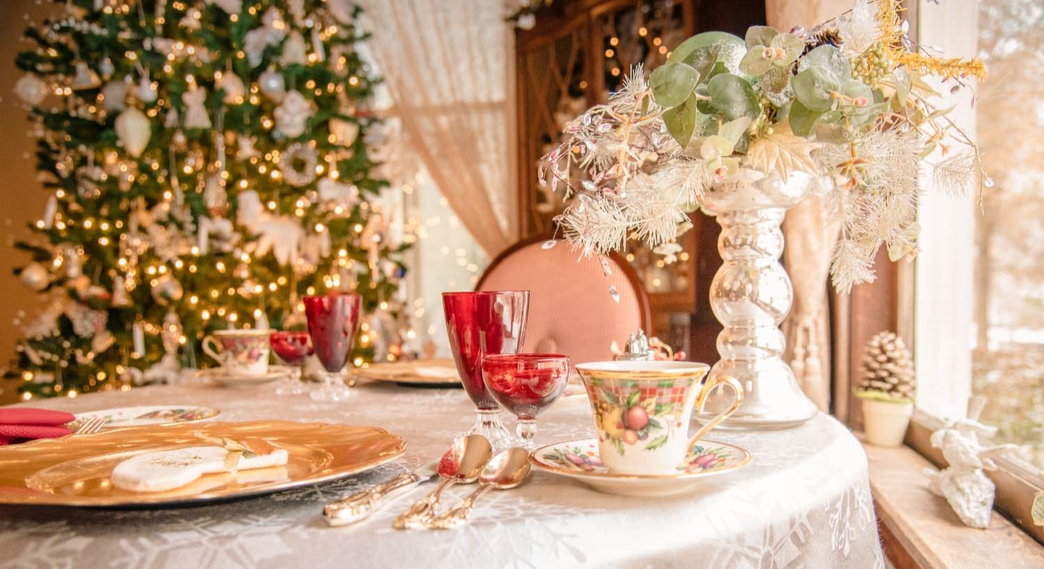Table set with fine china place settings, white snowflake tablecloth, and decorated Christmas tree in the background