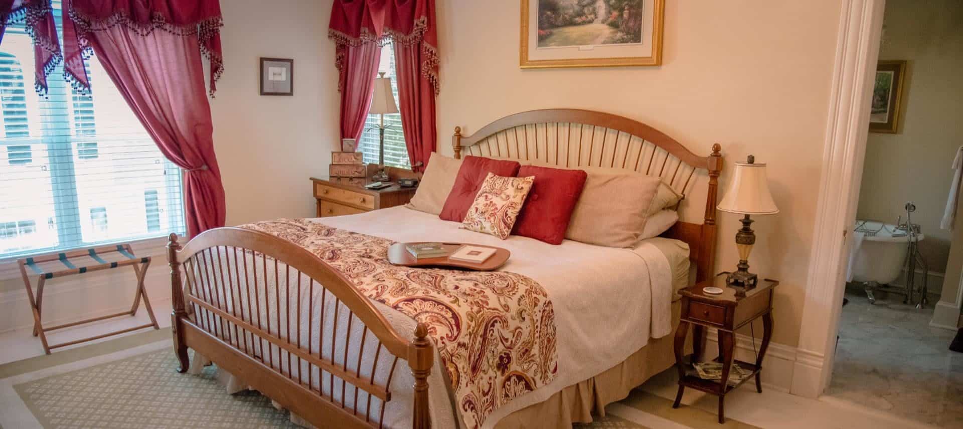 Bedroom with cream walls, wooden furniture and bed, white and tan bedding, and red curtains