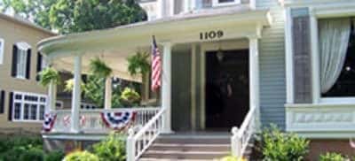 Front exterior view of the property painted blue and white with dark shutters, wrap around porch, American flags, and surrounded by green trees