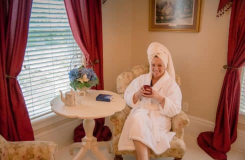 woman in fluffy white robe with white towel on her head sitting in a side chair with a mug of coffee